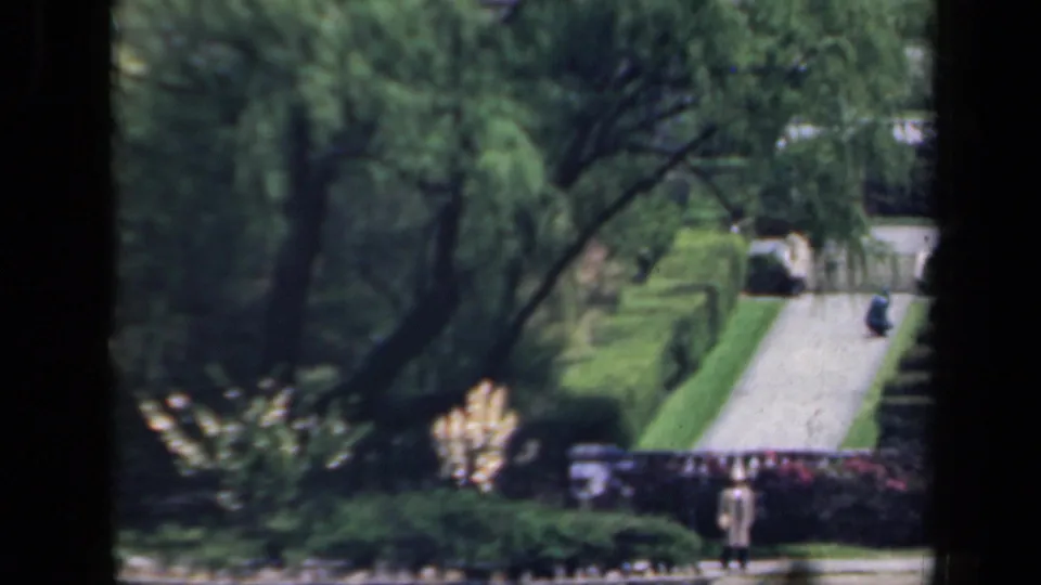 1941 New York 8mm frame with a distant garden visitor, trimmed hedges, trees, and a pale walkway in a Japanese-style garden setting.