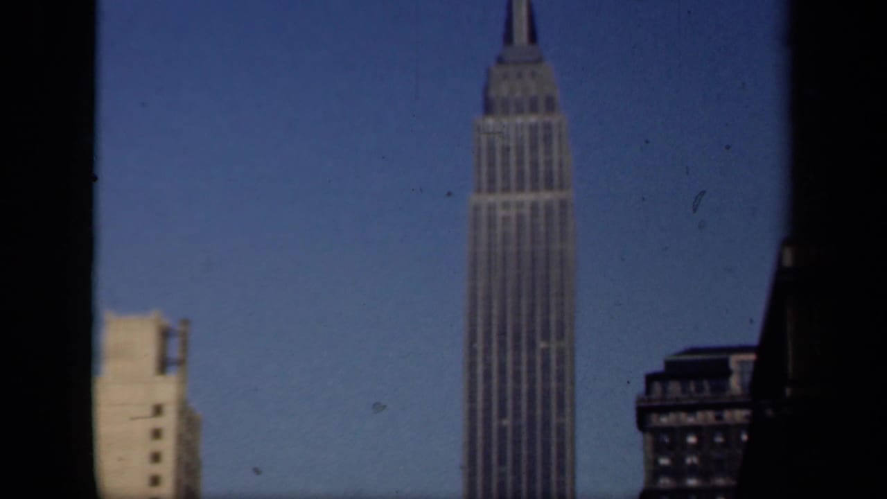 1941 New York home-movie frame with the Empire State Building and adjacent Midtown buildings under a clear blue sky.