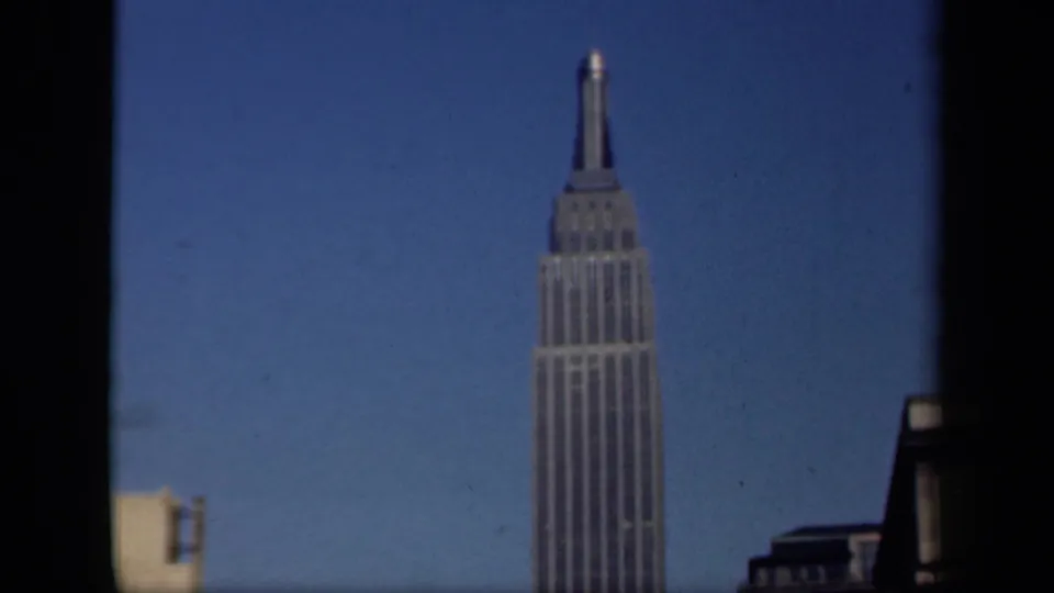 1941 New York 8mm frame of the Empire State Building seen through dark window edges, with neighboring rooftops in the foreground.