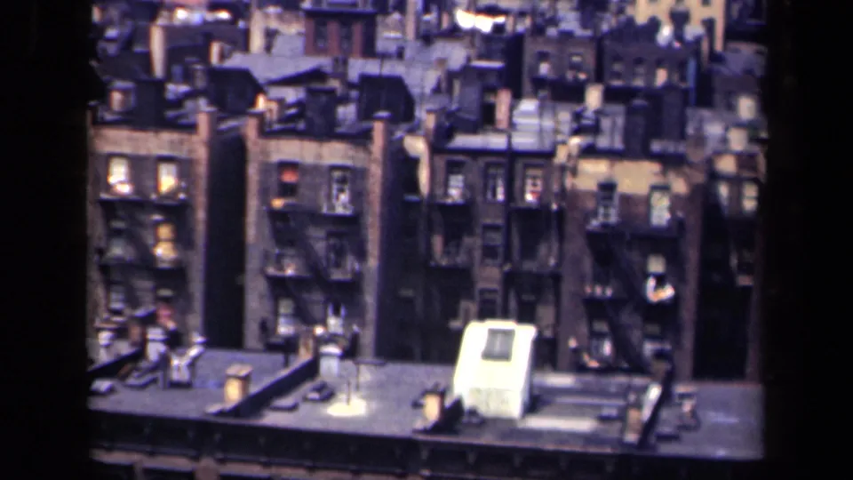 1941 New York home-movie frame showing dense apartment rows, rooftop structures, chimneys, and exterior fire escapes.