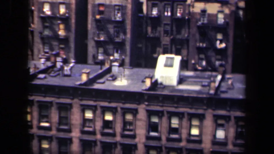 1941 New York city rooftop view with dense brick buildings, exterior fire escapes, vents and scattered illuminated windows.