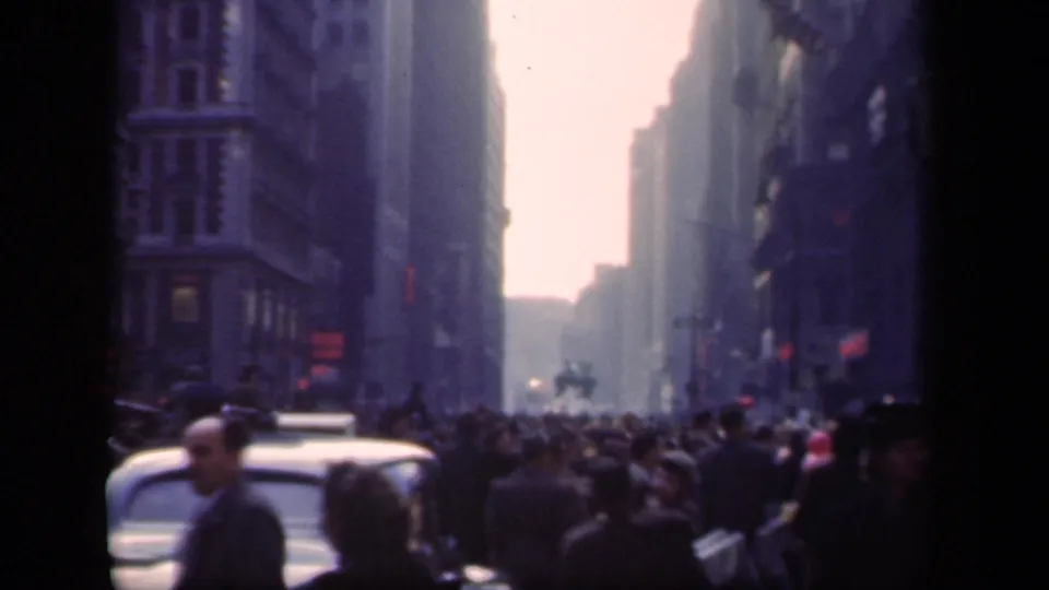 Soft 8mm frame of a crowded 1941 New York avenue with pedestrians, older automobiles, tall buildings, and traffic signals.