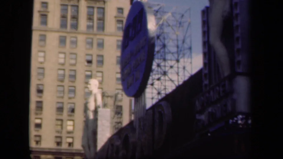 1941 New York home-movie frame showing a large blue sign, mannequin-like sidewalk display, metal billboard supports, and multi-story building windows.