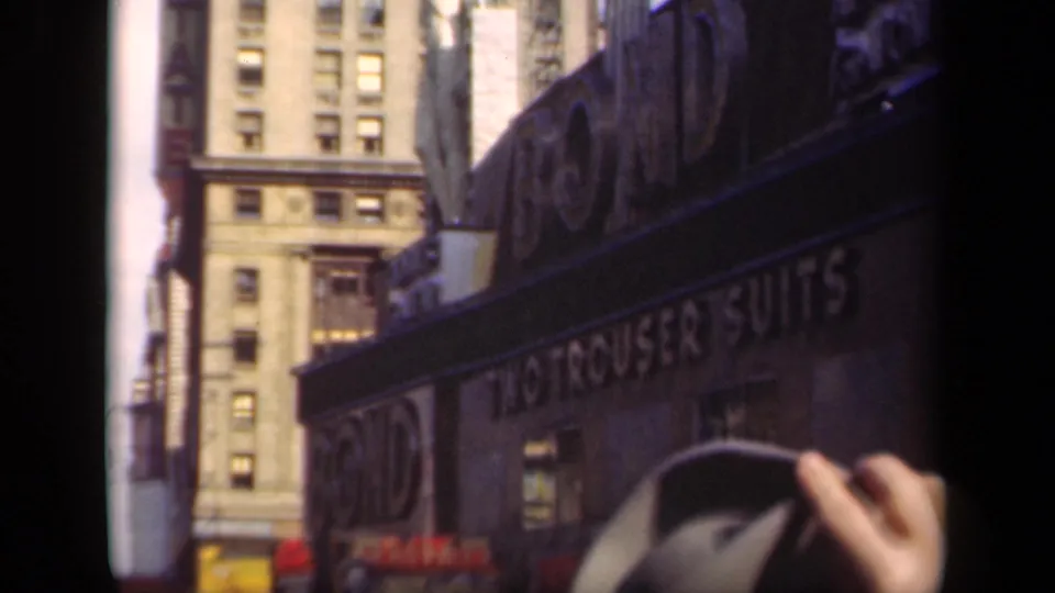 1941 New York storefront with visible two-trouser-suits lettering, dark apparel signage, and pedestrians beginning to enter the lower frame.