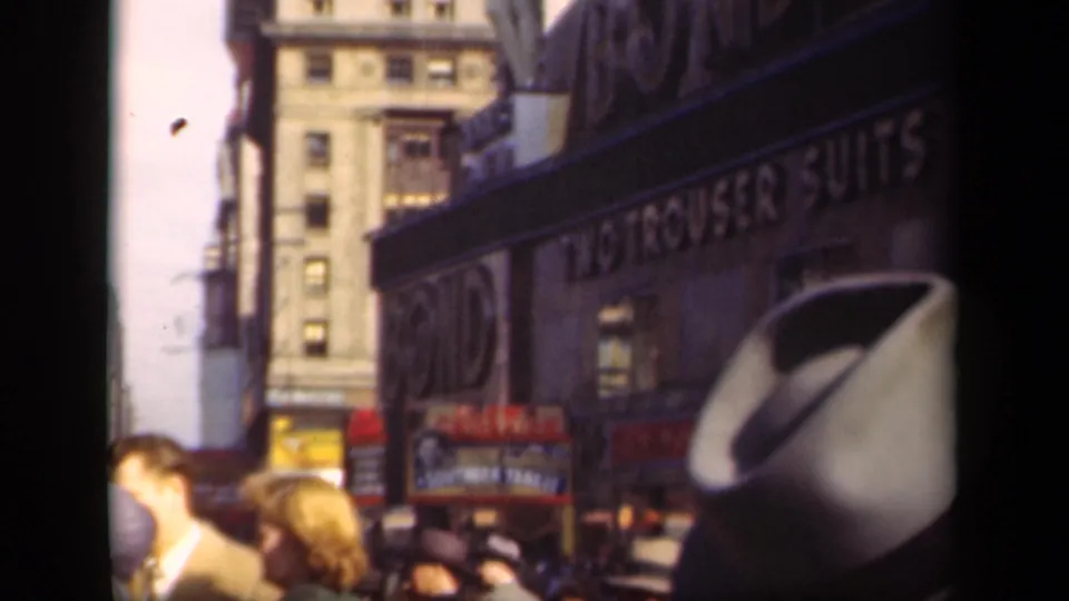 1941 New York street crowd moving below apparel and two-trouser-suits signs, with hats and faces visible in the foreground.
