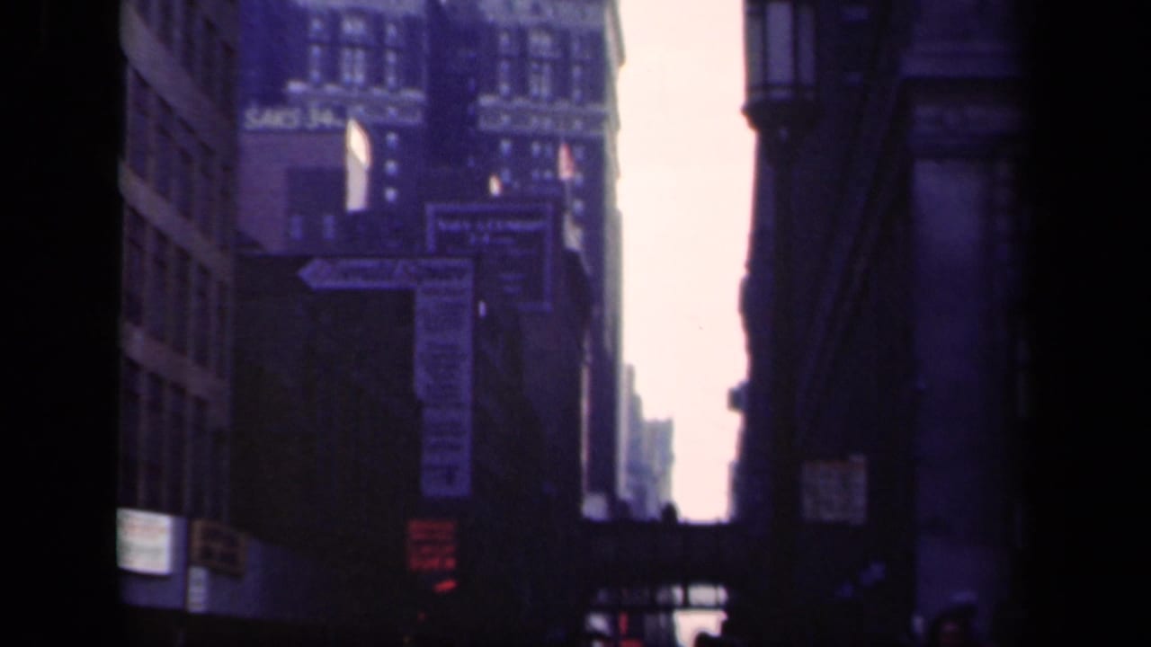 1941 New York street view showing shadowed building facades, commercial signage, and a tall downtown structure beyond.