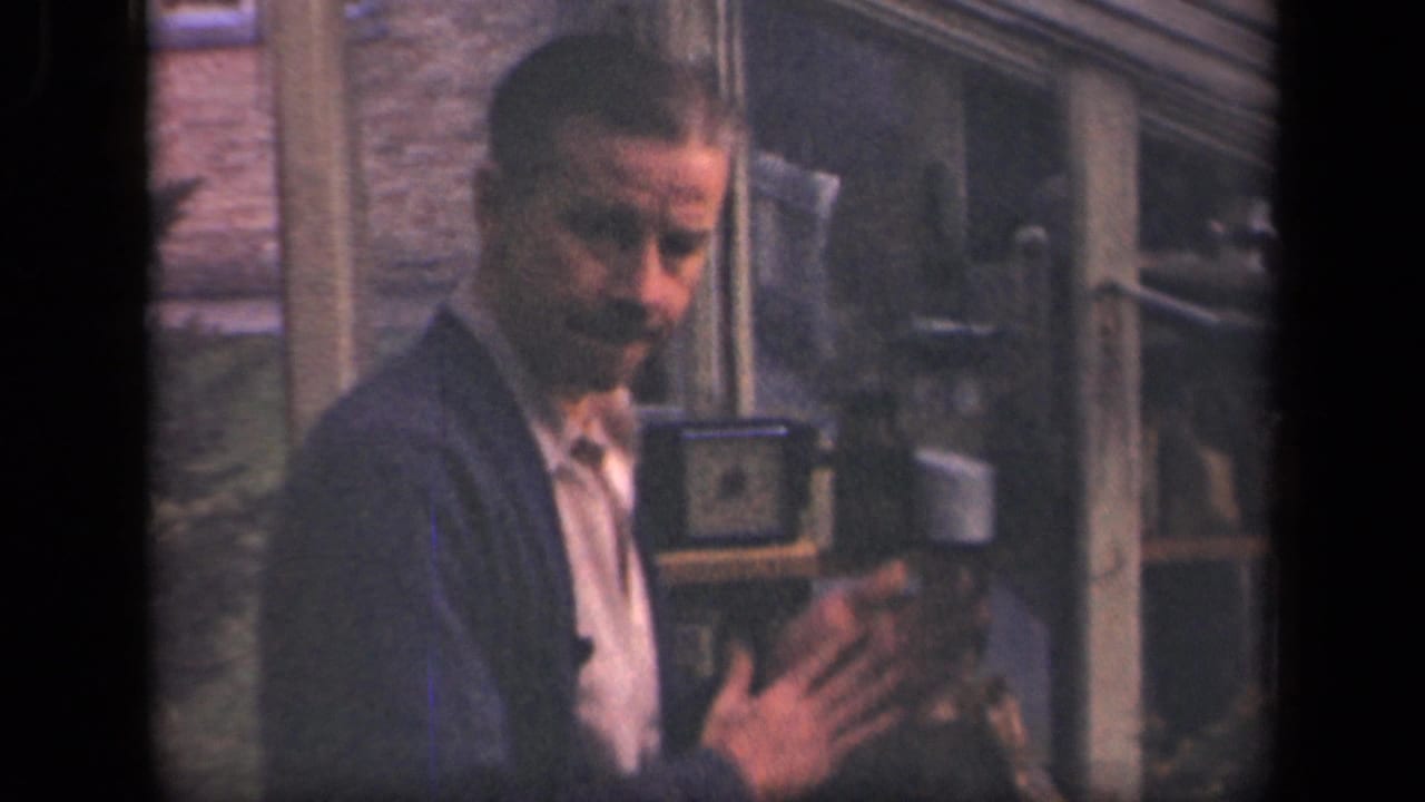In supported 1941 New York footage, a man in a cardigan stands in a lab-like interior with shelves, brick wall, and small apparatus.