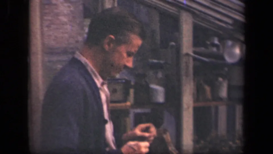A man in a cardigan looks down at a small object, with shelving and apparatus behind him in the New York 1941 footage.