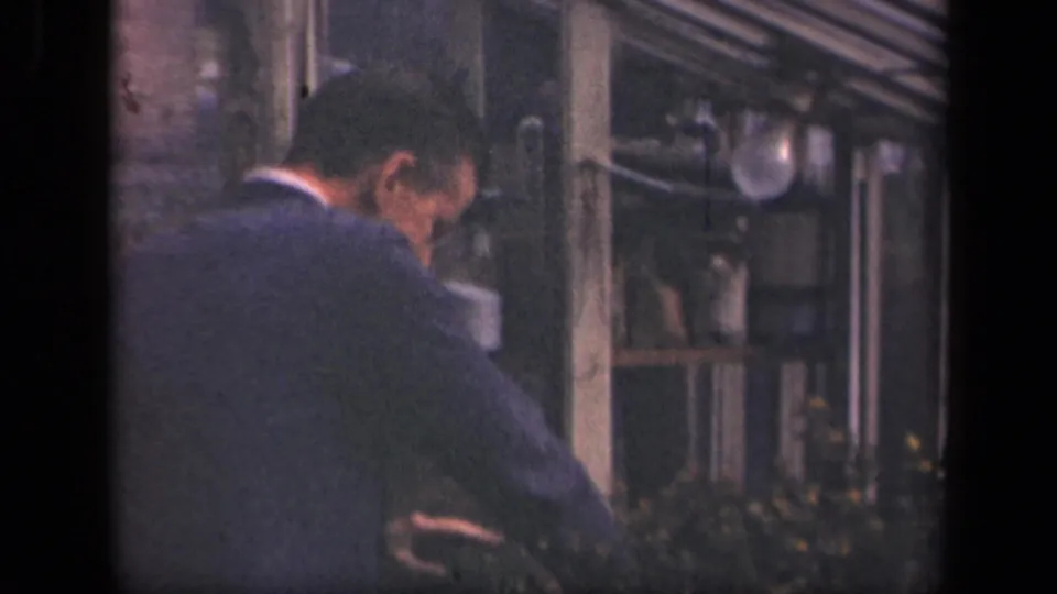 The man turns his back toward the camera while continuing work near shelves, glassware, and a window in 1941 New York footage.