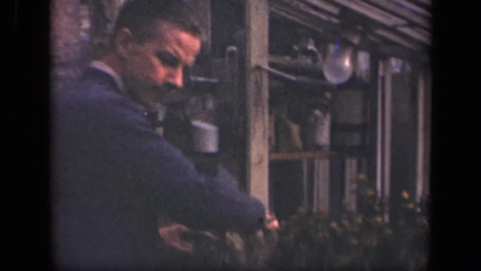 In 1941 New York home-movie footage, an adult man in a suit jacket bends toward shrubs beside windows and hanging pots.