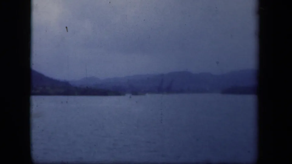 1941 8mm lake frame with a dark hillside, distant mountain ridge, and a small buoy-like mark on the water.
