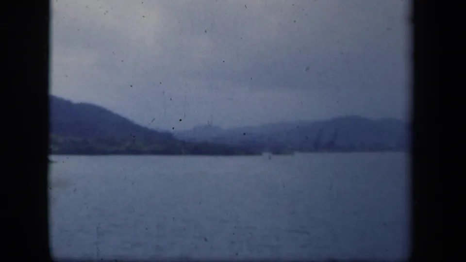 Blue-tinted 8mm frame of a New York lake scene, showing mountains, shoreline, and a small distant marker on the water.