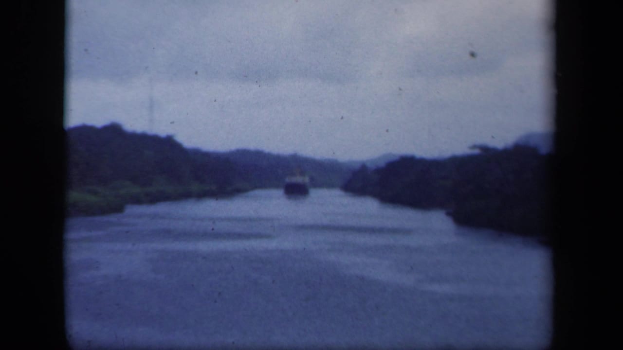 Distant ship on a narrow forested waterway under an overcast sky.