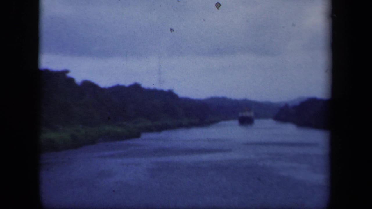 The boat remains distant on the New York waterway, with dark wooded banks and hazy hills visible through soft 8mm color.