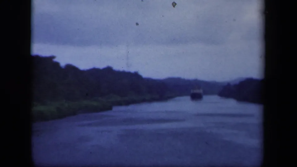 The boat remains distant on the New York waterway, with dark wooded banks and hazy hills visible through soft 8mm color.