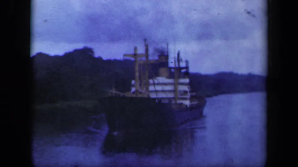 Blue-tinted 8mm view of a cargo ship navigating a calm, narrow waterway bordered by wooded hills in the 1941 New York clip.