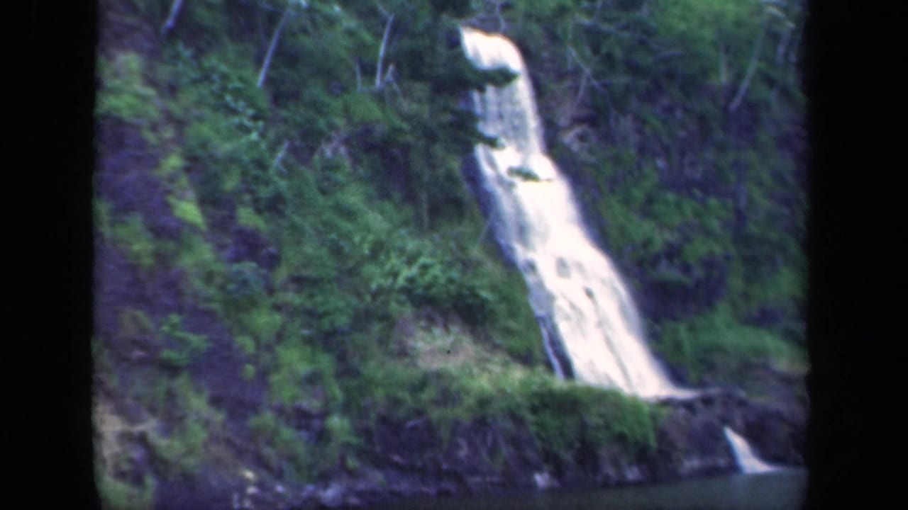 Vintage 8mm view of a New York waterfall running down dark rock through thick green forest, with soft exposure and frame edge shadows.