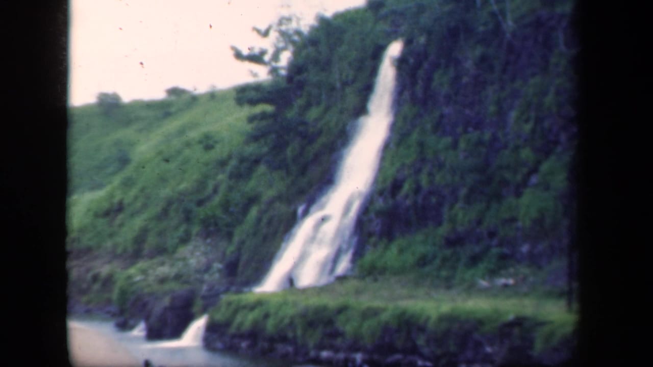 Waterfall dropping down a green hillside beside dark rock and water.