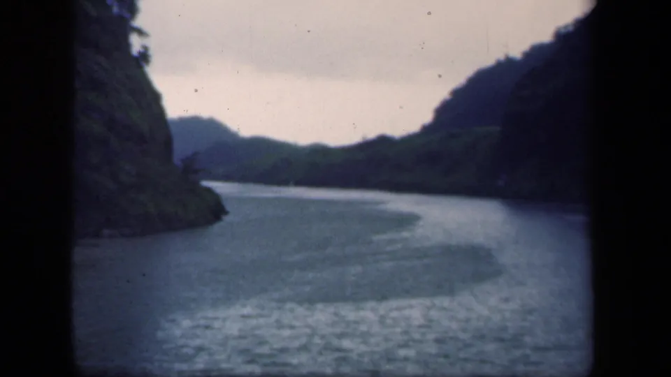 1941 New York home-movie frame of a river bend bordered by green banks and dark mountain slopes under an overcast sky.