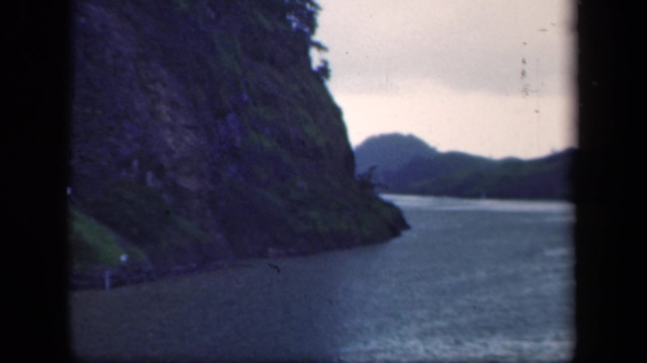 1940s 8mm frame of a steep green shoreline and calm waterway, filmed through dark vertical borders from a moving viewpoint.