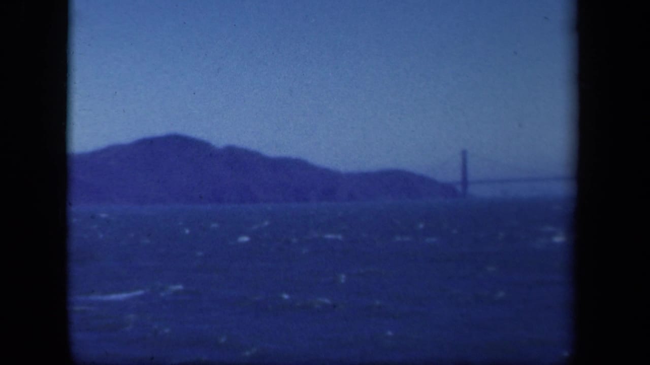 Distant view of the Golden Gate Bridge area across choppy bay water, with blue color shift and hills in the background.