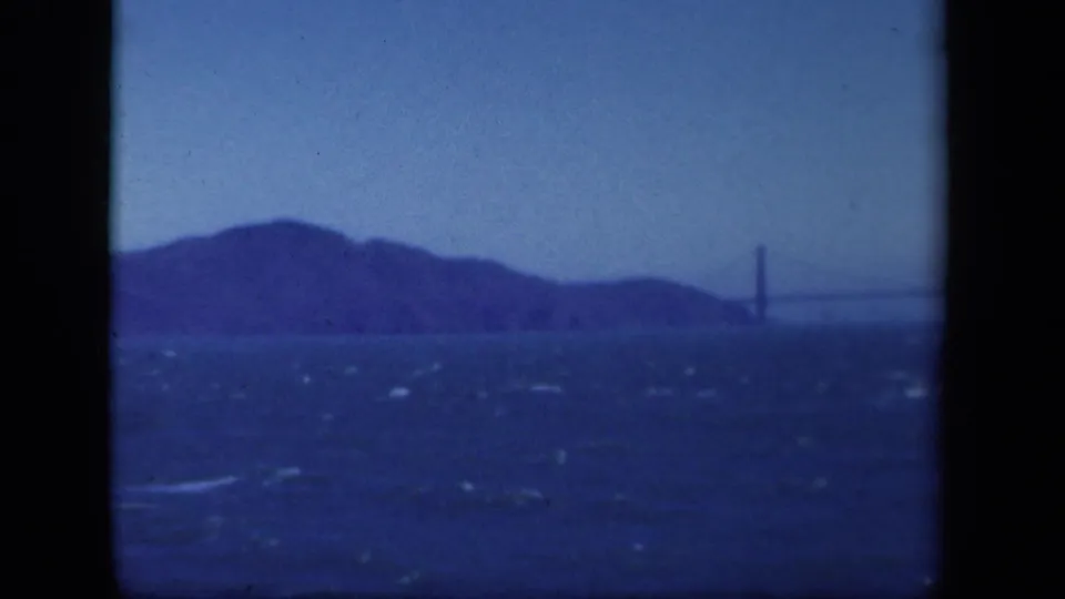 Distant view of the Golden Gate Bridge area across choppy bay water, with blue color shift and hills in the background.