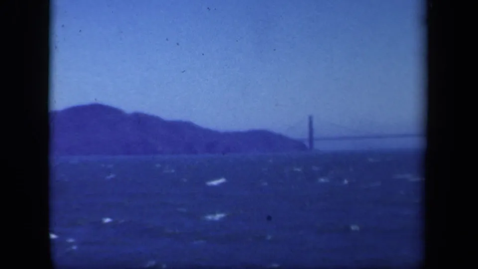 1940s 8mm bay view with distant hills, choppy water, and a suspension bridge partly visible near the right side of frame.