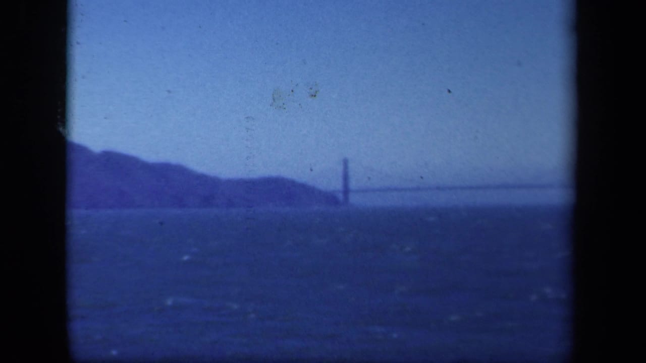 Distant suspension bridge across choppy water with low hills at left and dark film borders.
