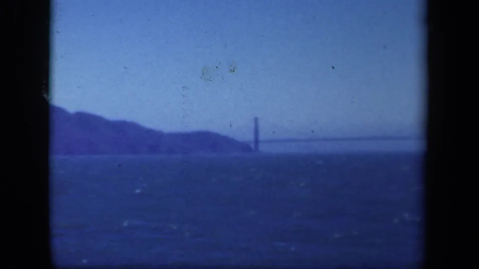 1941 8mm frame, metadata-located in New York, showing a distant suspension bridge across water with low shoreline hills and pale sky.