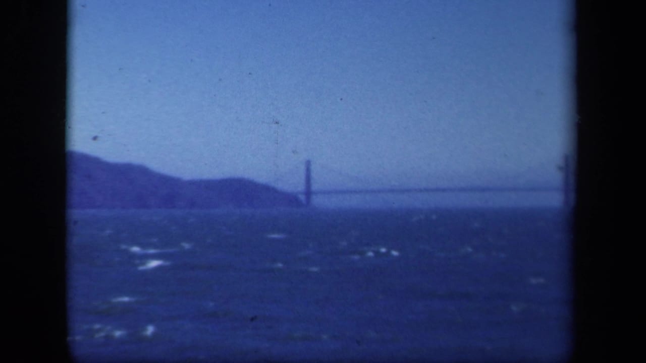 Soft blue 8mm view of a distant bridge crossing open water, with a tower near the left side and low hills behind it.