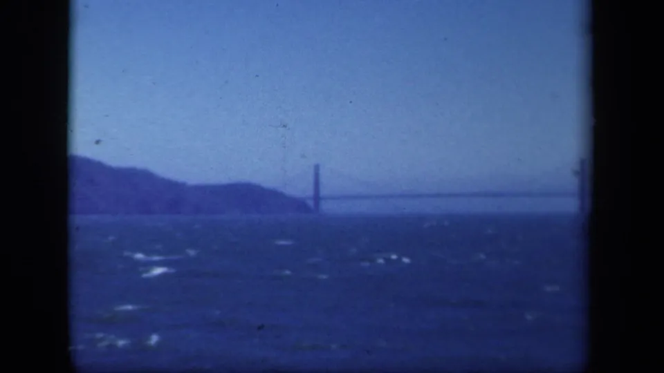 Soft blue 8mm view of a distant bridge crossing open water, with a tower near the left side and low hills behind it.
