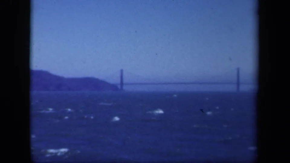 1941 home-movie frame of a suspension bridge in the distance, filmed over choppy water with shoreline hills at left.
