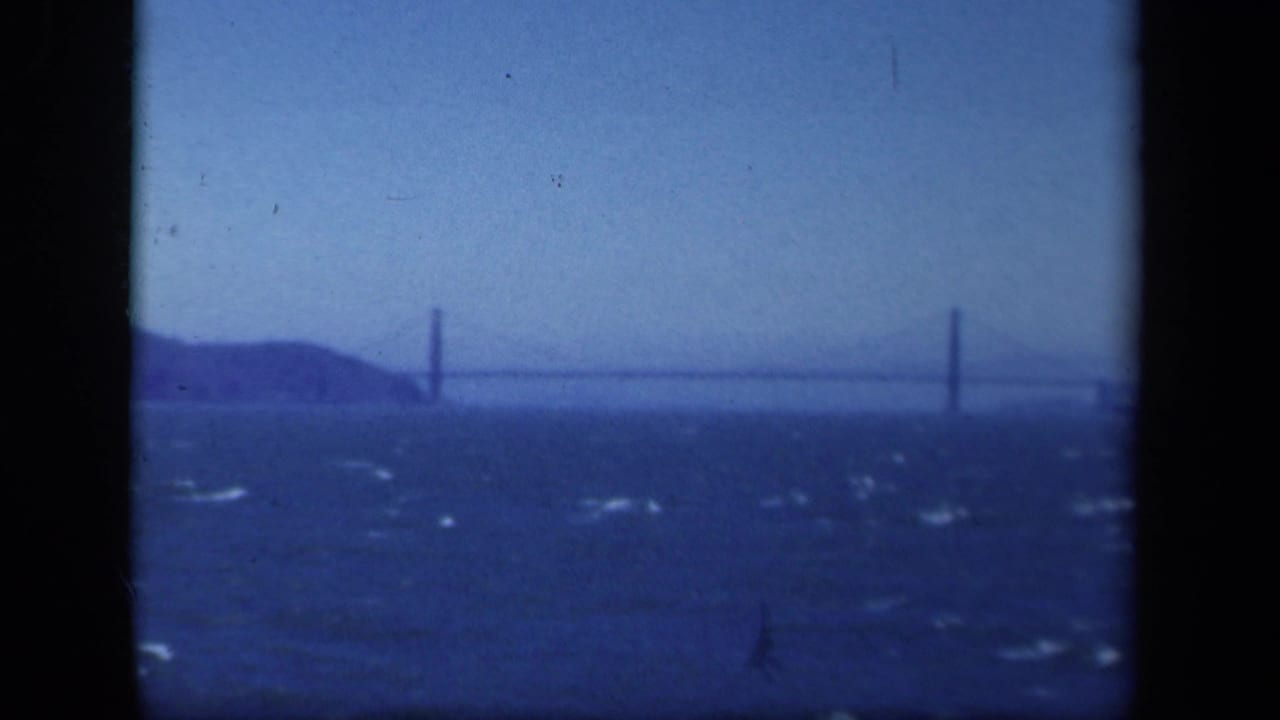 1941 New York-area 8mm frame showing a distant suspension bridge beyond dark, wind-ruffled water and a low shoreline.