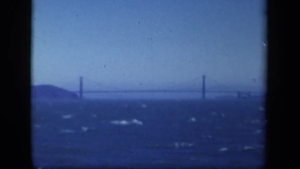 1941 New York-area home-movie frame with a faint bridge crossing the background behind rough blue water and dark gate edges.
