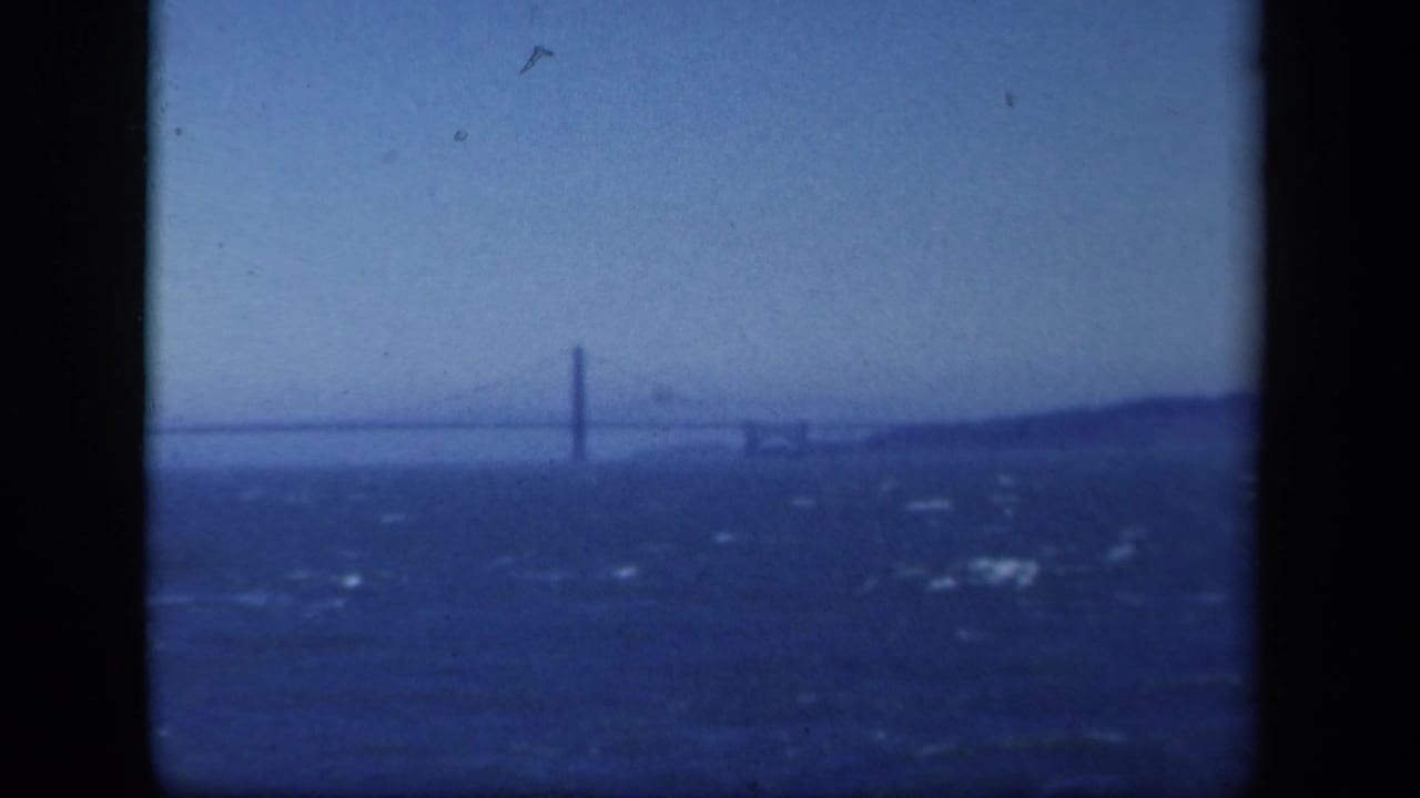 Distant 1941 New York bridge view with a tall tower, rough water in the foreground, and low shoreline behind it.