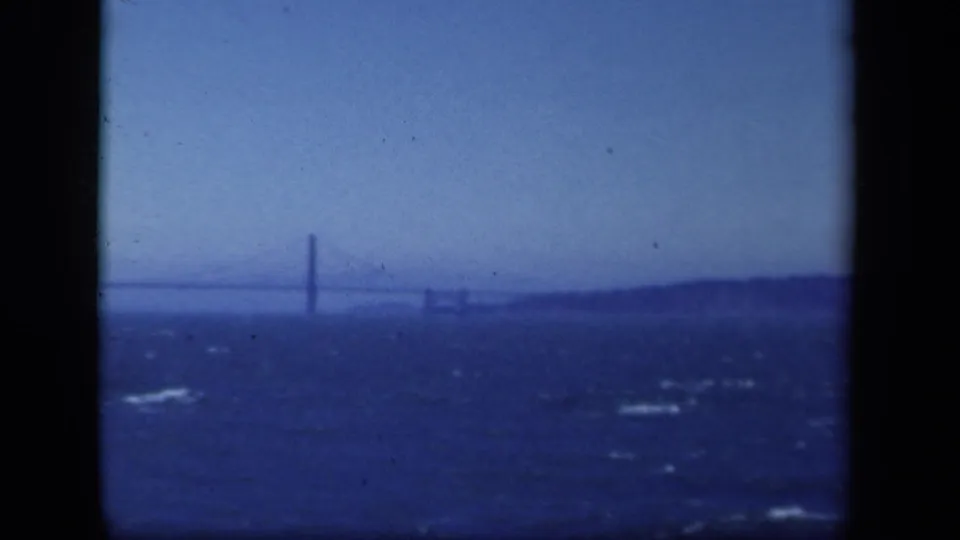 Soft 8mm frame of a distant New York suspension bridge, with waves and a hazy shoreline filling the view.