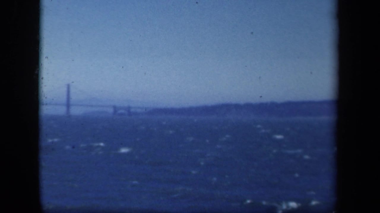 Distant view of a suspension bridge across bay water, likely filmed from a boat in archival 8mm footage.