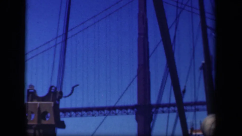 Suspension bridge deck and vertical cables appear behind ship hardware, with a blue-tinted 8mm film look.