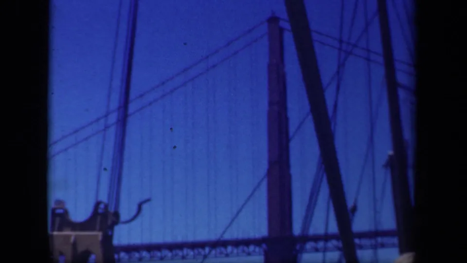 Large suspension bridge tower and cable pattern seen through boat rigging in blue-tinted archival 8mm footage.