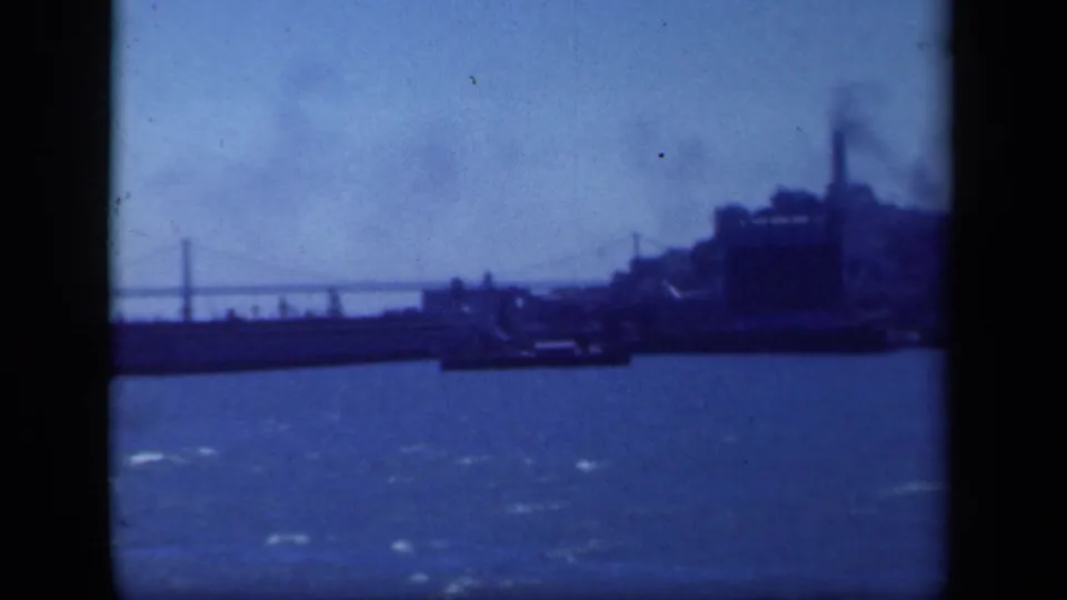 Soft 1941 home-movie frame of a waterfront scene, with open water, a distant bridge, shoreline structures, and smoke in the background.