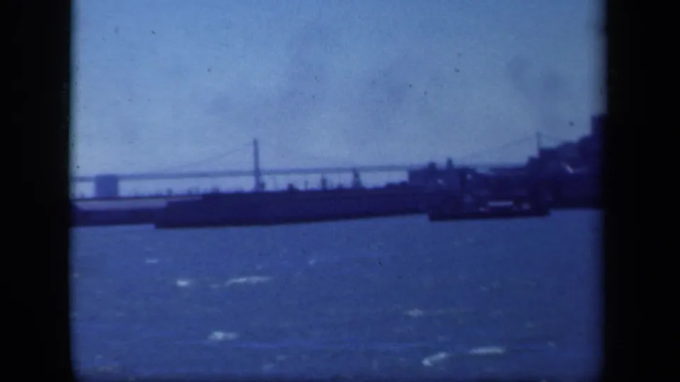 1941 New York-area home-movie frame with a suspension bridge beyond blue water and a dark low shoreline or vessel form.