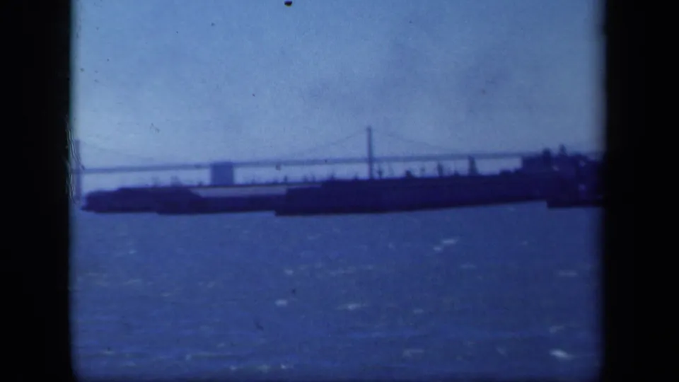 Soft 8mm frame of a New York-area waterfront panorama, with bridge cables above a long dark vessel or pier-like form.