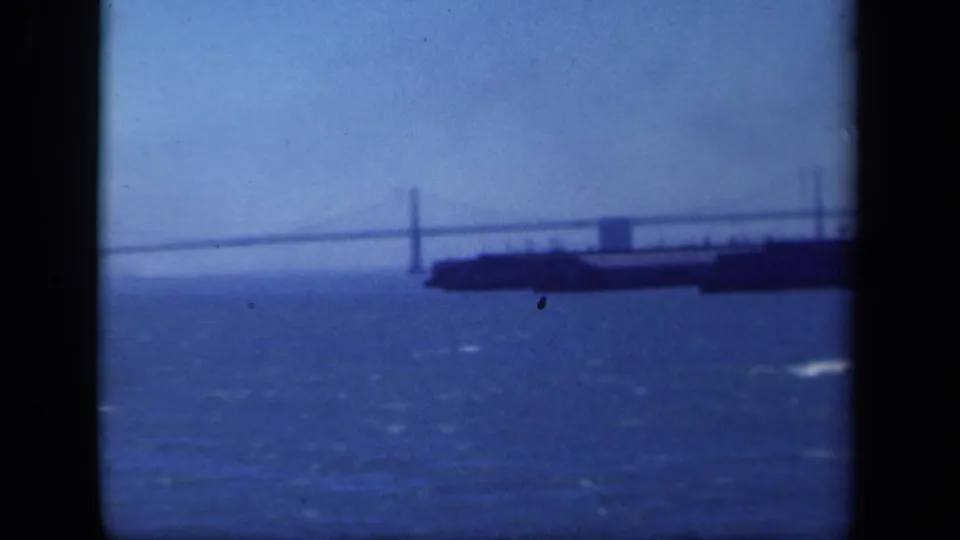 1940s 8mm harbor view showing a distant suspension bridge, choppy water, and a dark ship or barge near the span.