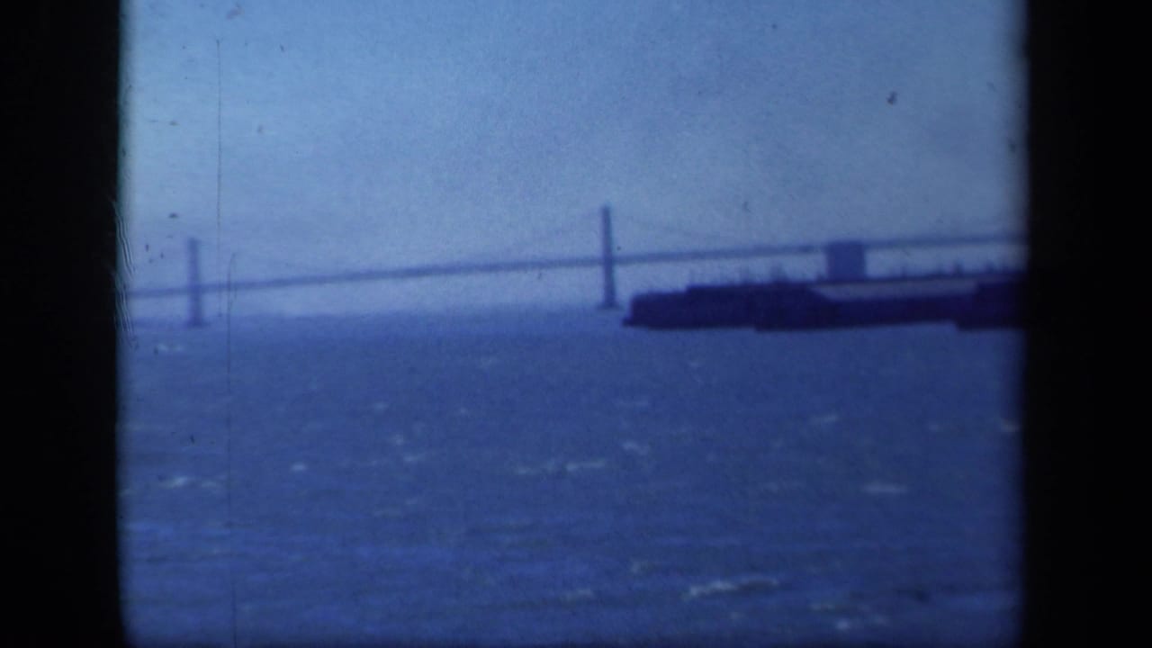 Blue-tinted 8mm frame of a suspension bridge across a harbor, with a large vessel near the right edge and choppy water below.