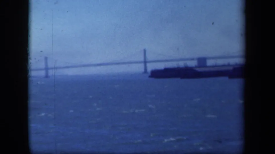 Blue-tinted 8mm frame of a suspension bridge across a harbor, with a large vessel near the right edge and choppy water below.