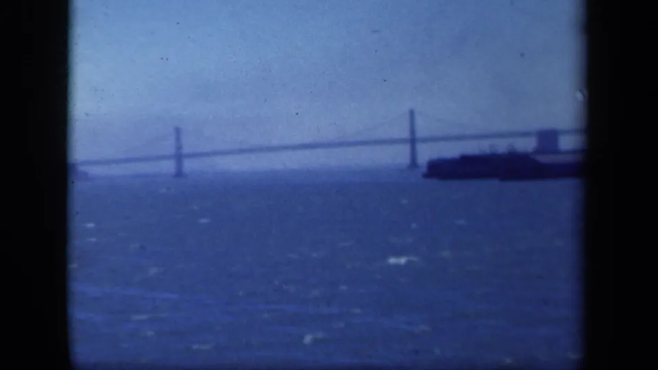 Distant 1940s waterfront view with a suspension bridge crossing the frame and a dark vessel partly visible against the water.
