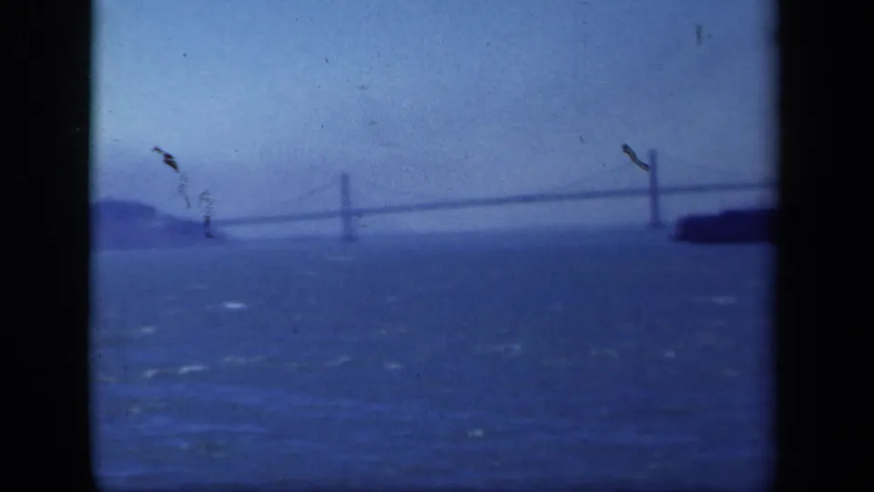 Final 8mm frame showing a hazy suspension bridge over open water, distant landform, and the edge of a passing vessel.