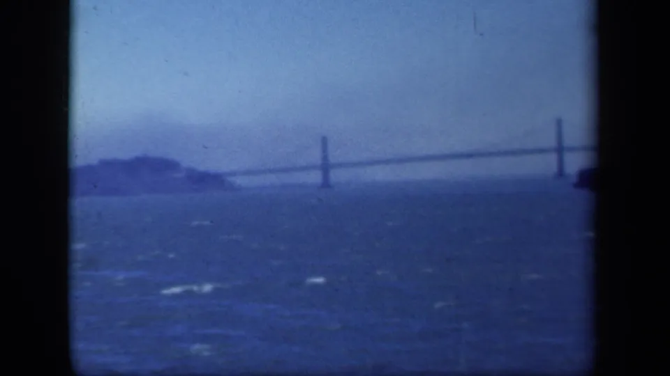 1941 8mm home-movie frame showing choppy bay water, a distant island, and a long suspension bridge under a hazy blue sky.
