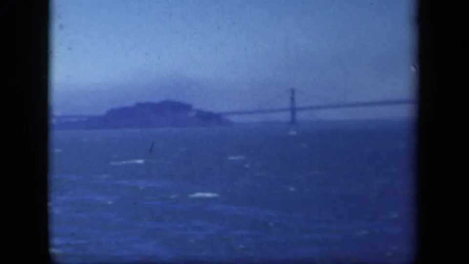 Blue-tinted 8mm frame of a long suspension bridge crossing behind a small island, filmed from water in the listed 1941 location.