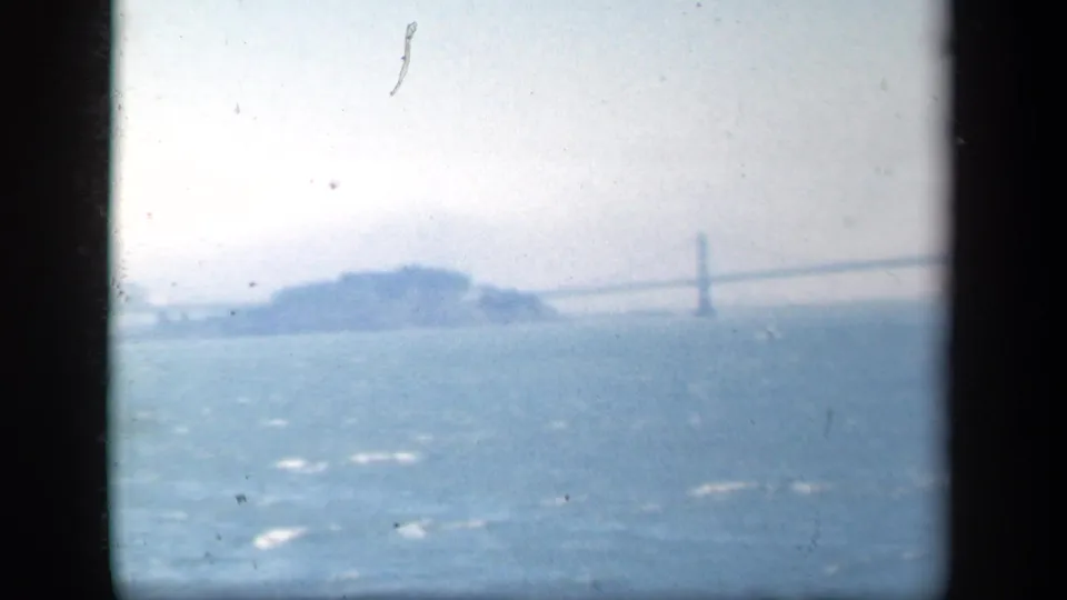 Washed, bright 8mm frame showing pale water, a distant island, and a faint suspension bridge partly softened by exposure and haze.